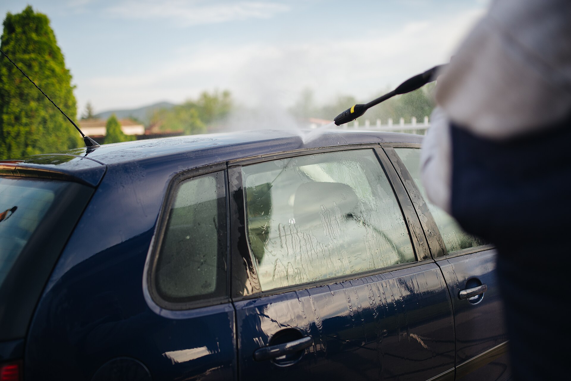 Pressure washing a blue car in a sunny residential driveway